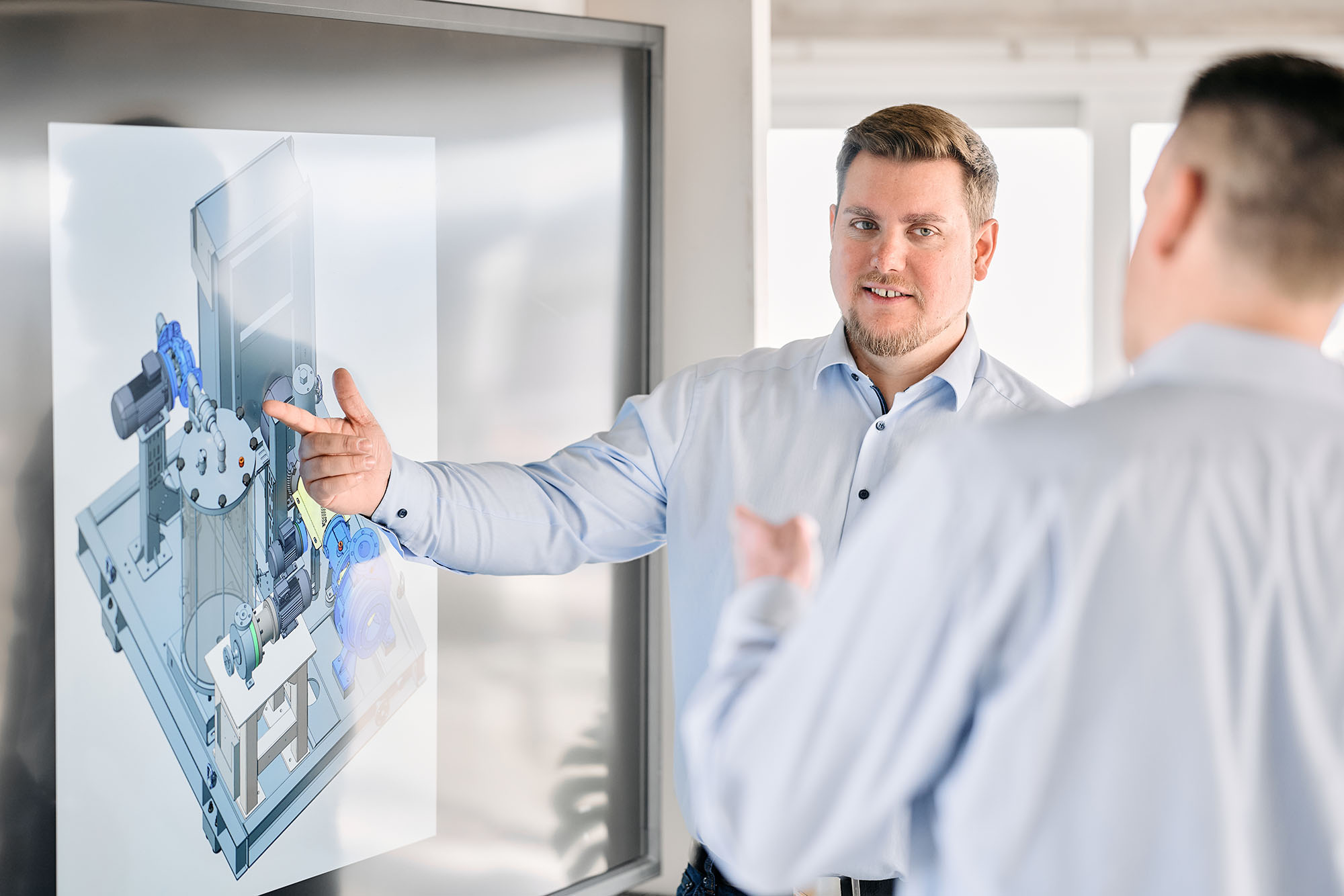 Home 2 Mitarbeiter erklärt einem Kollegen eine technische Zeichnung auf einem Großbildschirm, Businessfotografie Köln